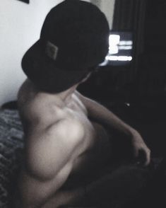 a shirtless man sitting on top of a bed wearing a black hat and no shirt