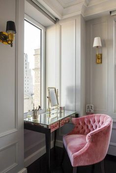 a pink chair sitting in front of a window next to a desk with a mirror on it