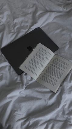 an open book laying on top of a bed next to an apple laptop computer monitor