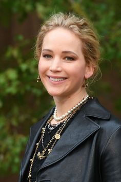 a woman in a black leather jacket and pearls smiles at the camera with trees behind her