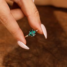 a woman's hand holding a ring with green stones on it and white nails