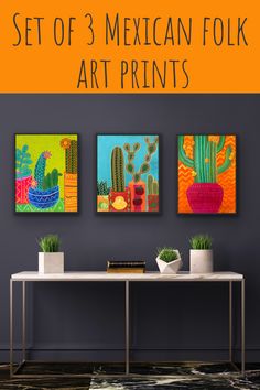 three mexican folk art prints hanging on the wall above a table with two potted cacti