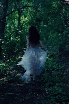 a woman is walking through the woods with her dress blowing in the wind and trees behind her