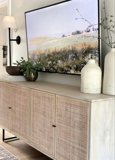 a white vase sitting on top of a wooden cabinet next to a painting and lamp