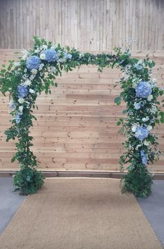 an outdoor wedding arch with blue flowers and greenery on the top, in front of a wooden wall