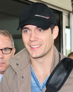 a man wearing a cap and glasses smiles at the camera while standing next to another man