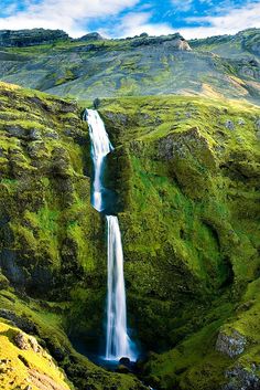 a large waterfall in the middle of a green mountain with grass on it's sides