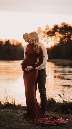 a pregnant couple standing next to each other in front of a body of water at sunset