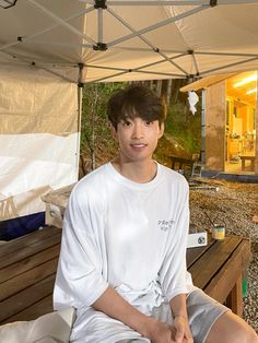 a young man sitting on top of a wooden bench next to a white tarp