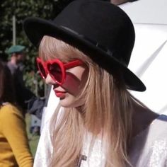 a woman with long blonde hair wearing red sunglasses and a black hat, standing in front of a white tent