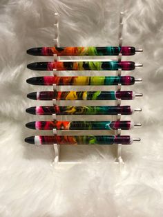 six pens are lined up in a row on a white furnishing area, one is multicolored and the other is black