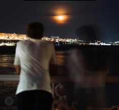 two people looking out over the water at night with full moon in background and city lights