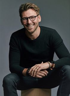 a man with glasses sitting on top of a wooden stool smiling at the camera while wearing a black sweater and jeans