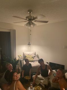 four people are sitting on the floor in a room with a bed and ceiling fan