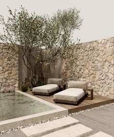 two lounge chairs sitting next to a pool in front of a stone wall and tree