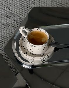 a coffee cup with saucer and spoon sitting on a glass table top next to a cell phone