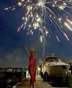 a woman in a red dress is standing on a dock with her arms up as fireworks go off