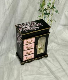 a pink and black jewelry box next to a plant on a white cloth covered background