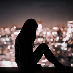 a woman sitting in front of a window with the city lights behind her at night