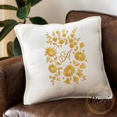 a white pillow with gold monogrammed flowers and the letter d on it sitting on a brown leather chair