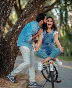 a man and woman kissing on the back of a bike in front of some trees