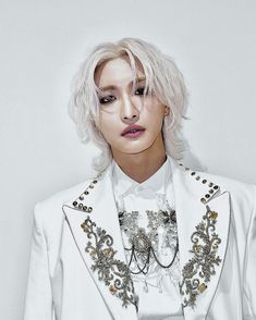 a woman with white hair wearing a white jacket and silver jewelry on her neck, standing in front of a white wall