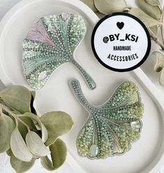 two decorative items on a white plate next to some green plants and leaves with the words bylsi handmade accessories