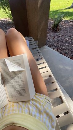 a person laying down reading a book on a bench in the shade with their legs crossed