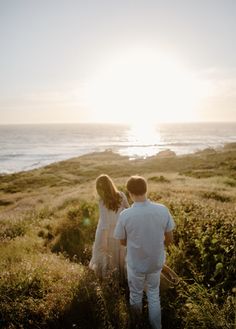 a man and woman walking down a hill towards the ocean with the sun behind them