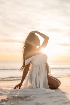 a woman sitting on top of a sandy beach next to the ocean in white dress