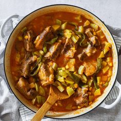 a pot filled with meat and vegetables on top of a white table cloth next to a wooden spoon