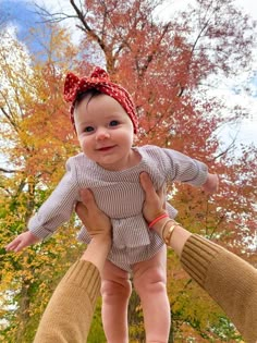 a woman holding a baby in her arms with autumn trees in the back ground behind her