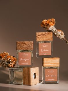 three small square jars with labels on them sitting next to dried flowers and leaves in front of a gray background