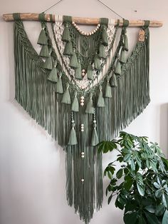 a green wall hanging with tassels and beads on it next to a potted plant