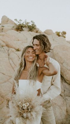 two women standing next to each other in front of some rocks and grass with one woman holding her arm around the man's shoulder