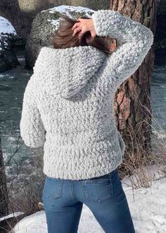 a woman standing next to a tree in the snow wearing a white sweater and jeans