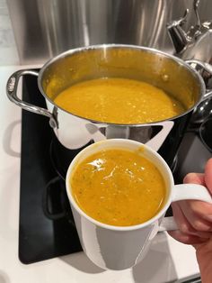 a person is holding a cup of soup in front of a pot on the stove