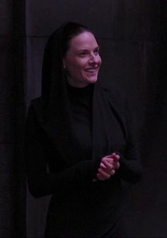 a woman standing in front of a black wall with her hands together and smiling at the camera