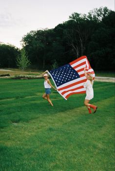 two girls running with an american flag on the grass in front of some trees and bushes