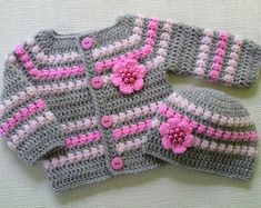 two crocheted sweaters with pink flowers on them and one has a matching hat