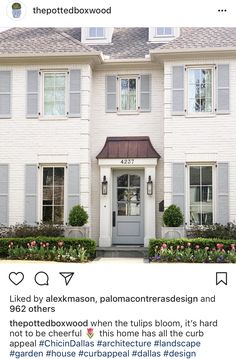 a white house with gray shutters on the front and side doors, surrounded by flowers