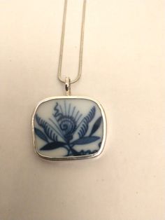 a blue and white square pendant with flowers on it's side, hanging from a silver chain