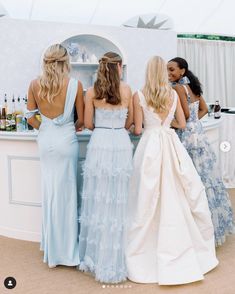 three women in dresses are standing at a bar with bottles on the counter and one woman is wearing a white dress