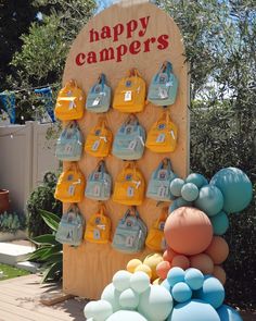 A large wooden slab with signage atop that says "Happy Campers" in red letters with yellow and blue backpacks in multiple mini sizes hanging from it and various balloons in size and color at the base Summer Camp Theme Birthday Party, Explorer Birthday Theme, Camper Party Theme, Camping One Year Birthday, Campground Birthday Party, Camp Party Decor, Camp Birthday Party Adult, 70s Camping Aesthetic, Camping Theme Birthday Party Decorations