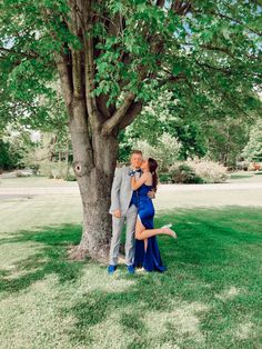 a man and woman standing next to a tree in the grass with their arms around each other