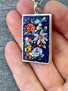 a hand holding a small square pendant with flowers on it's side and blue glass in the middle