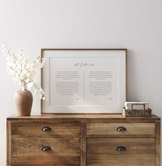 a vase with flowers on top of a wooden dresser next to a white wall and framed poem