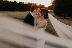 a bride and groom kissing in the middle of a field at sunset with veil blowing over them