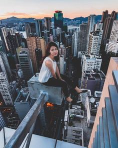 a woman sitting on the edge of a high rise overlooking cityscape at sunset