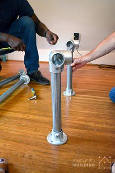 two men working on an electrical device in a room with hard wood flooring and exposed pipes